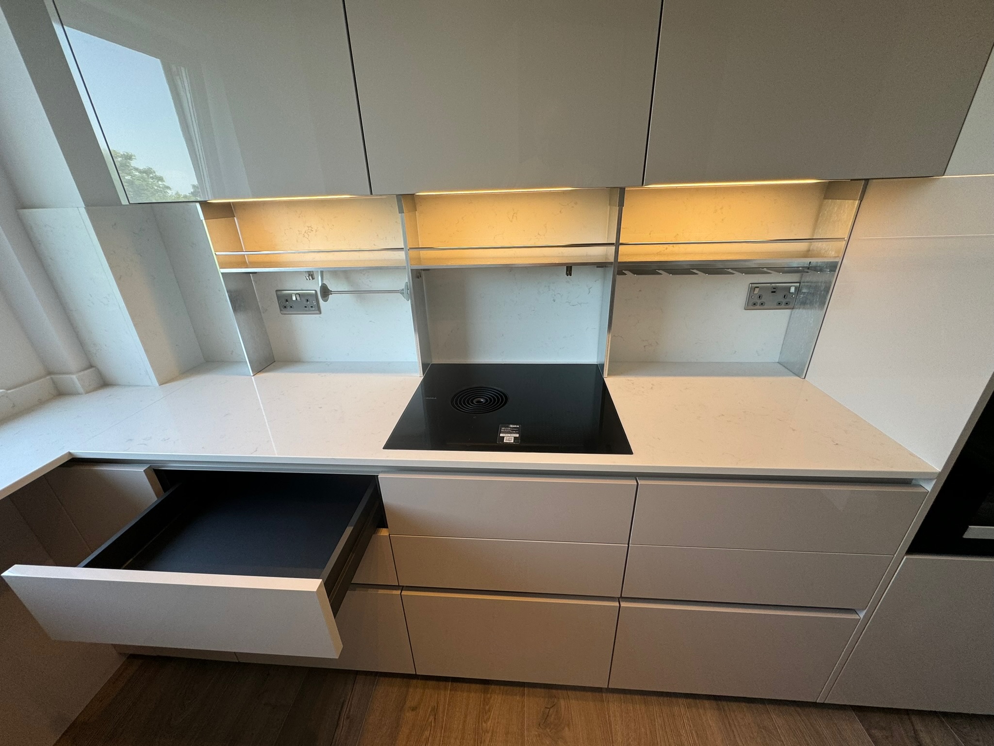 Pale handleless kitchen with an open storage drawer, integrated extractor hob and illuminated wall storage